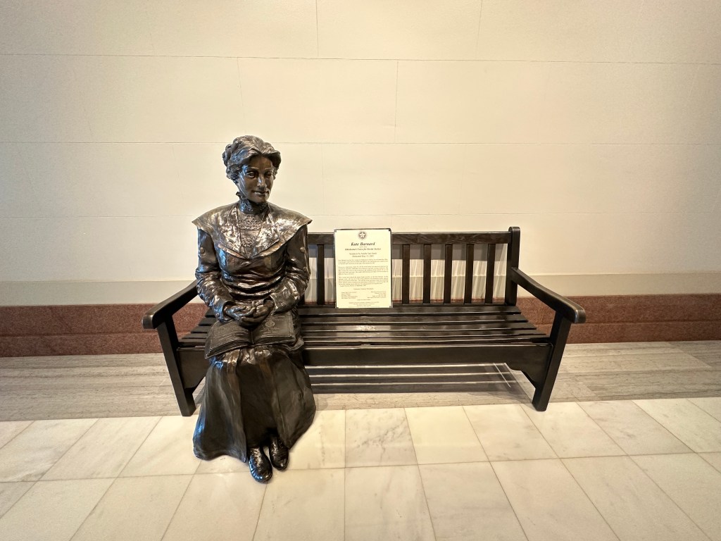 Bronze statue of a woman sitting on a bench, commemorating significant contributions, with a plaque beside her detailing her achievements.