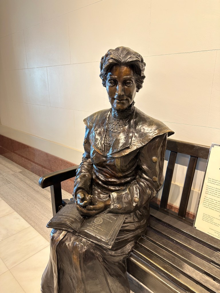 Bronze statue of a seated woman holding a book, depicted in a historical costume, placed on a bench in a light-colored interior.