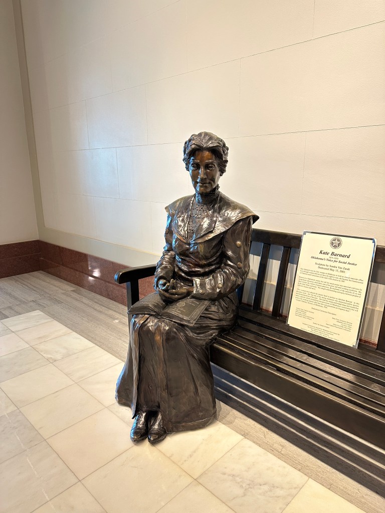 Bronze statue of a seated woman wearing a historical dress, holding a book, displayed on a bench in an indoor setting.