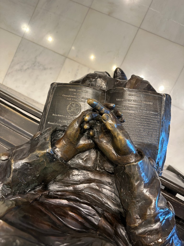 Close-up view of a bronze sculpture depicting a person reading a book, with hands clasped together over the pages, showcasing intricate details.