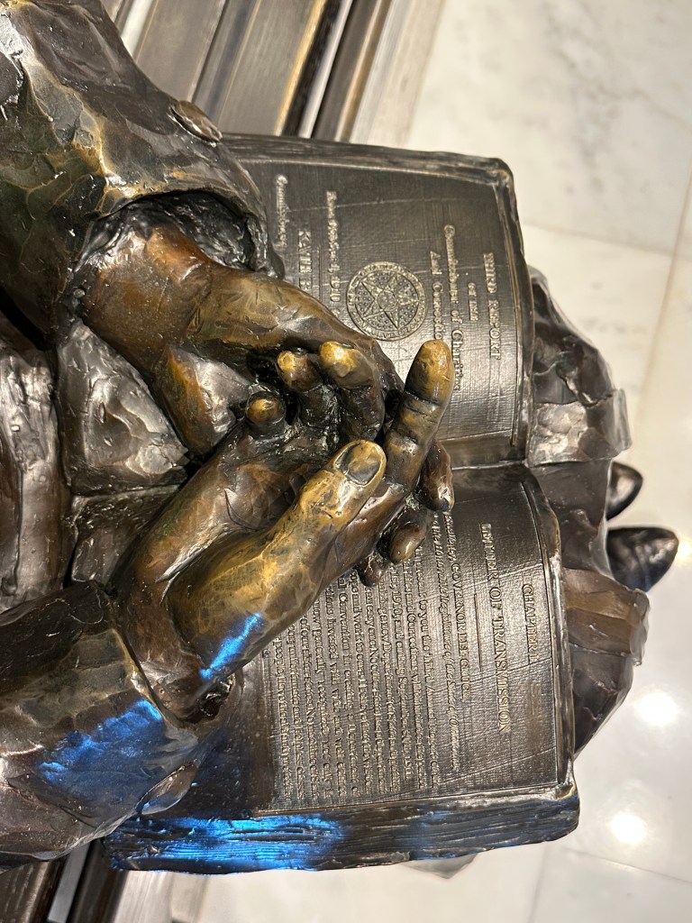 Close-up view of a bronze sculpture depicting hands clasped over a book, with text visible on the book's pages.