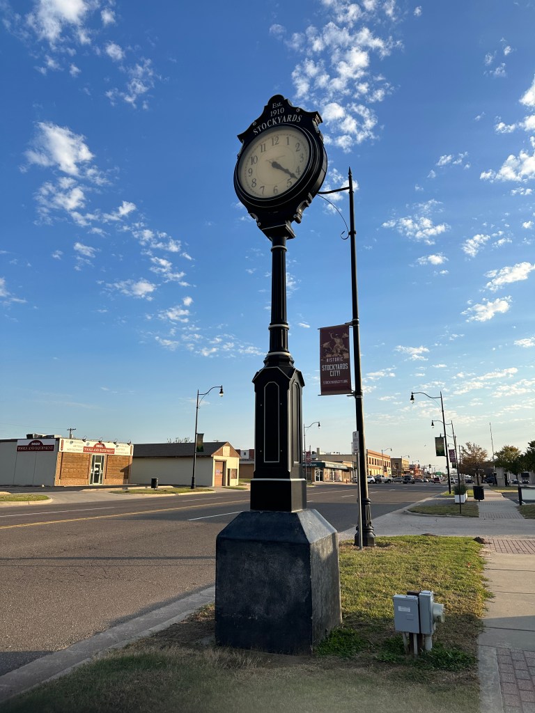 A traditional street clock with 'Stockyards' and 'Est. 1910' engraved at the top, positioned on a sidewalk with a clear blue sky in the background.