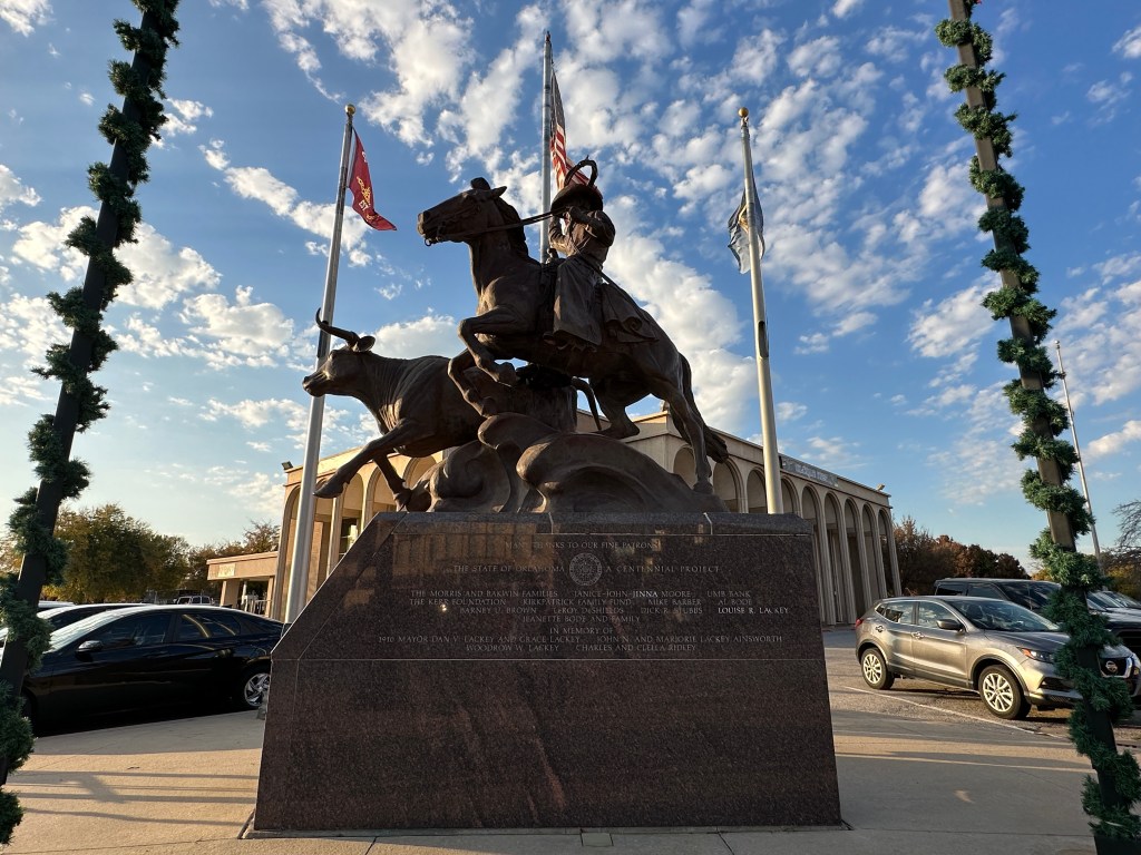 A bronze statue of a cowboy on horseback in front of a building, with flags flying in the background and decorative greenery nearby.