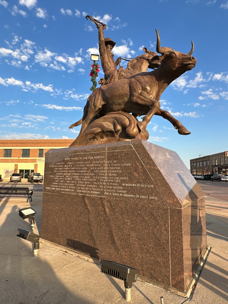 A bronze sculpture of a cowboy on horseback, holding a rope, with a bull in front. The statue is mounted on a granite base featuring inscriptions and sponsor acknowledgments. The background showcases a blue sky with clouds.