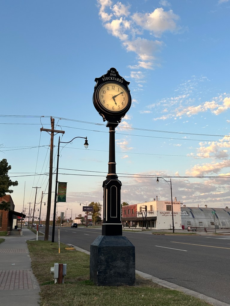A vintage street clock with the inscription 'Est. 1910 Stockyards' stands on a sidewalk beside a quiet street, with a clear blue sky and scattered clouds in the background.