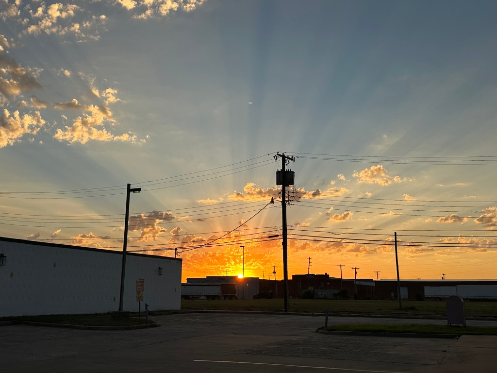 Sunset with vibrant orange and yellow rays behind clouds, silhouetted power lines and buildings in the foreground.