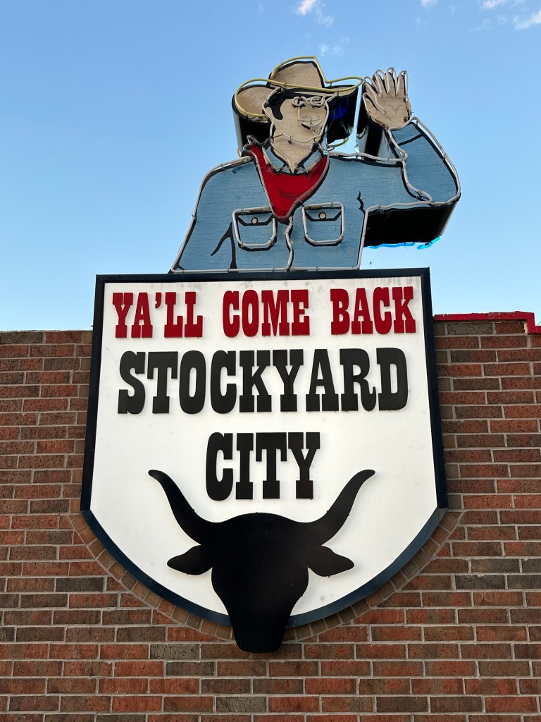 Sign for Stockyard City featuring a cowboy figure waving and the phrase 'Ya'll come back'.