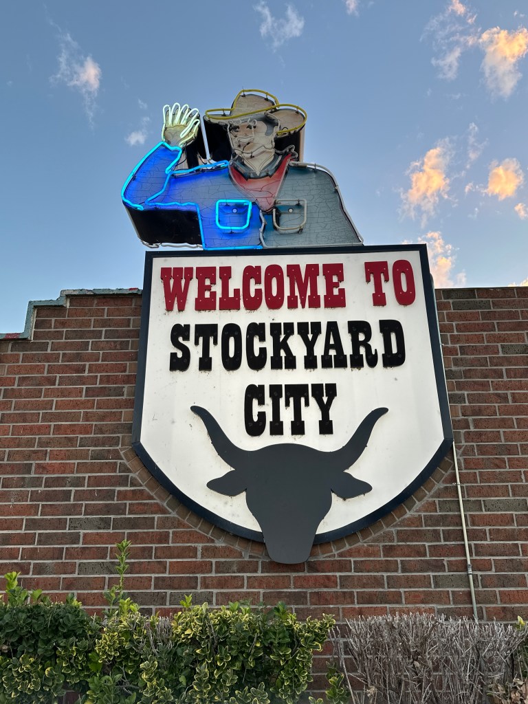 A welcome sign for Stockyard City featuring a cowboy figure waving and a longhorn silhouette, set against a brick background with a colorful sky.