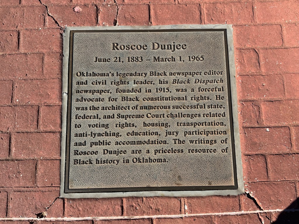Bronze historical marker plaque detailing the achievements of Roscoe Dunjee, an influential Black newspaper editor and civil rights leader in Oklahoma, set against a red brick background.