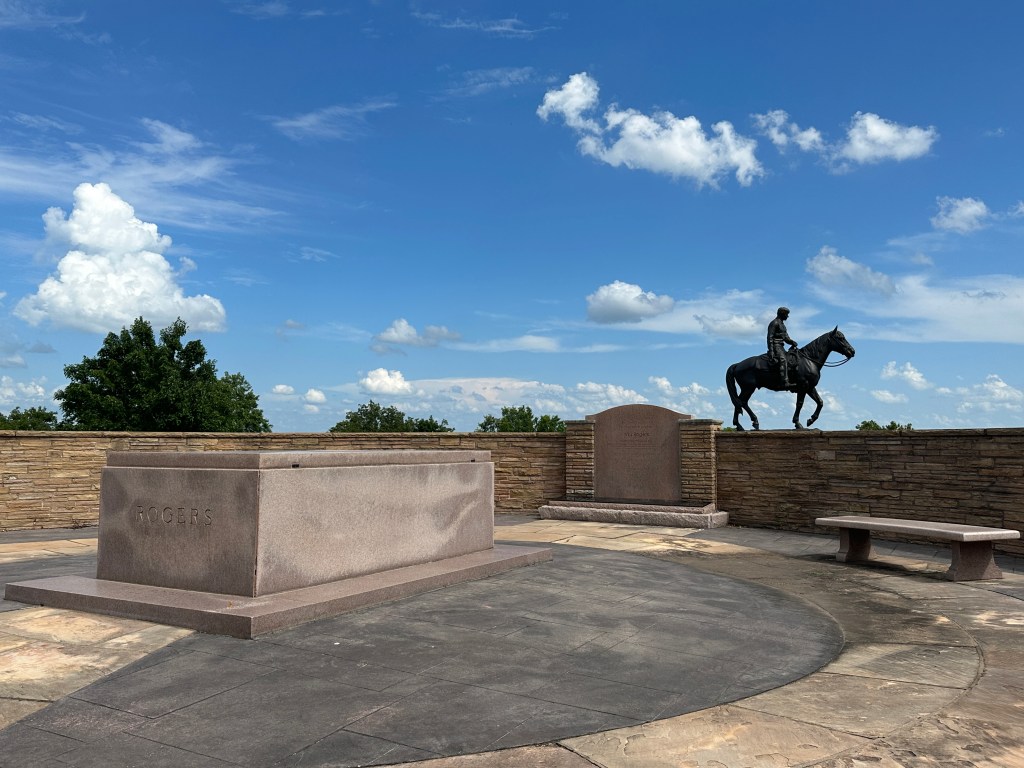 The Will Rogers Memorial site featuring a horse statue and gravestone surrounded by a stone wall under a blue sky with clouds.