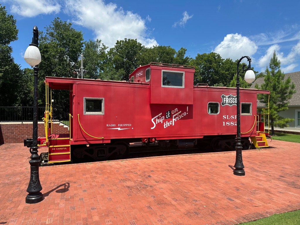 Red train caboose displayed outside with signs reading 'Ship it on the Frisco!' and 'Radio Equipped', surrounded by green trees and blue sky.