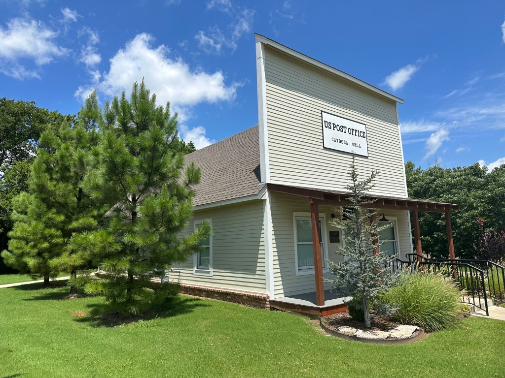 Exterior view of the replica of the original US Post Office in Catoosa, Oklahoma, surrounded by greenery and under a blue sky.