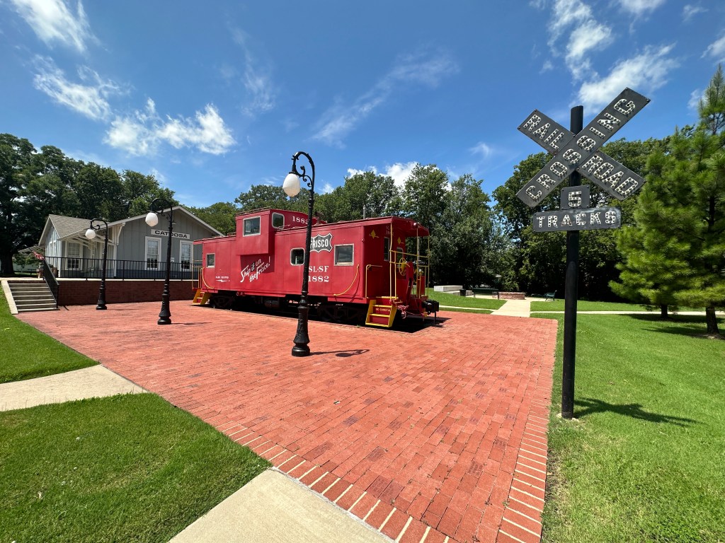 A red train caboose stands prominently on a brick pathway in front of a building labeled 'Catoosa.' The scene includes green grass, a railroad crossing sign, and decorative street lamps under a blue sky.