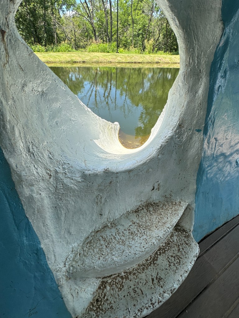 Interior view of a hollowed-out section of the Blue Whale in Catoosa, Oklahoma, showing a round opening overlooking a calm pond with lush greenery in the background.