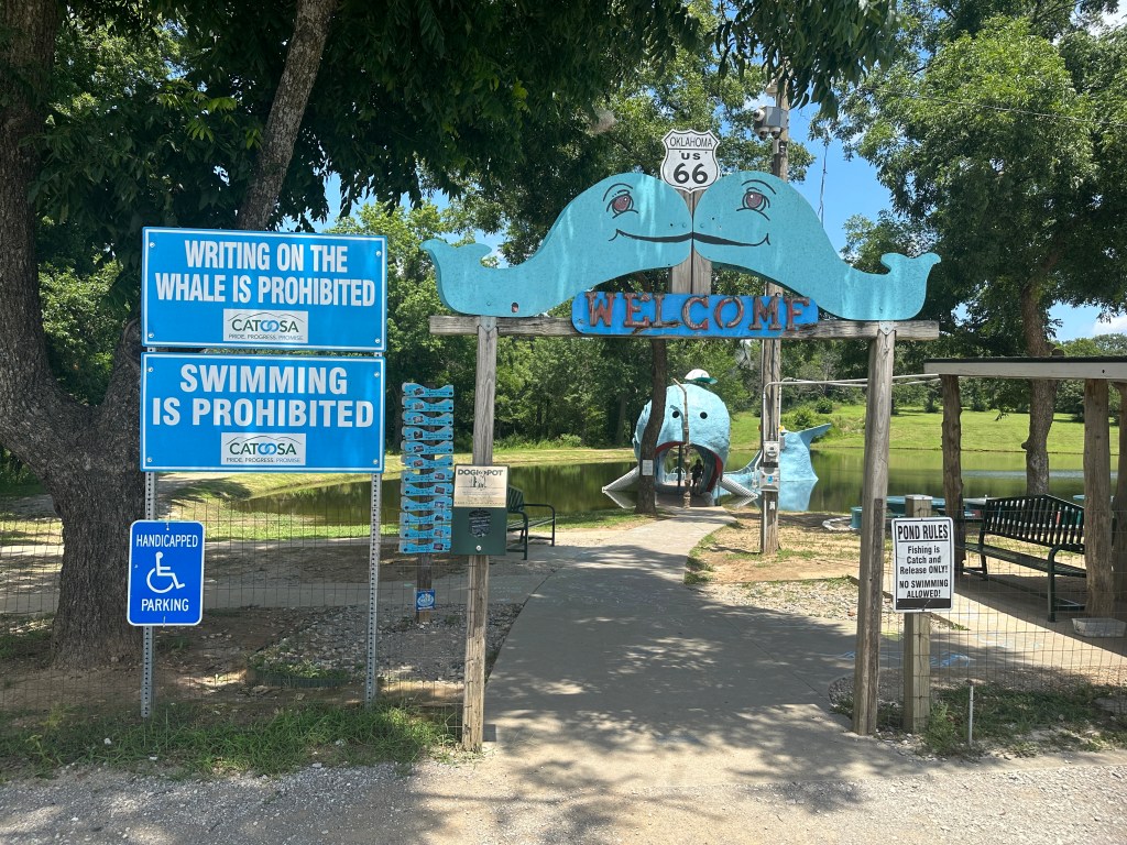 Entrance to the Blue Whale in Catoosa, Oklahoma, featuring signs prohibiting swimming and writing on the whale, along with a welcome sign and a view of the pond.