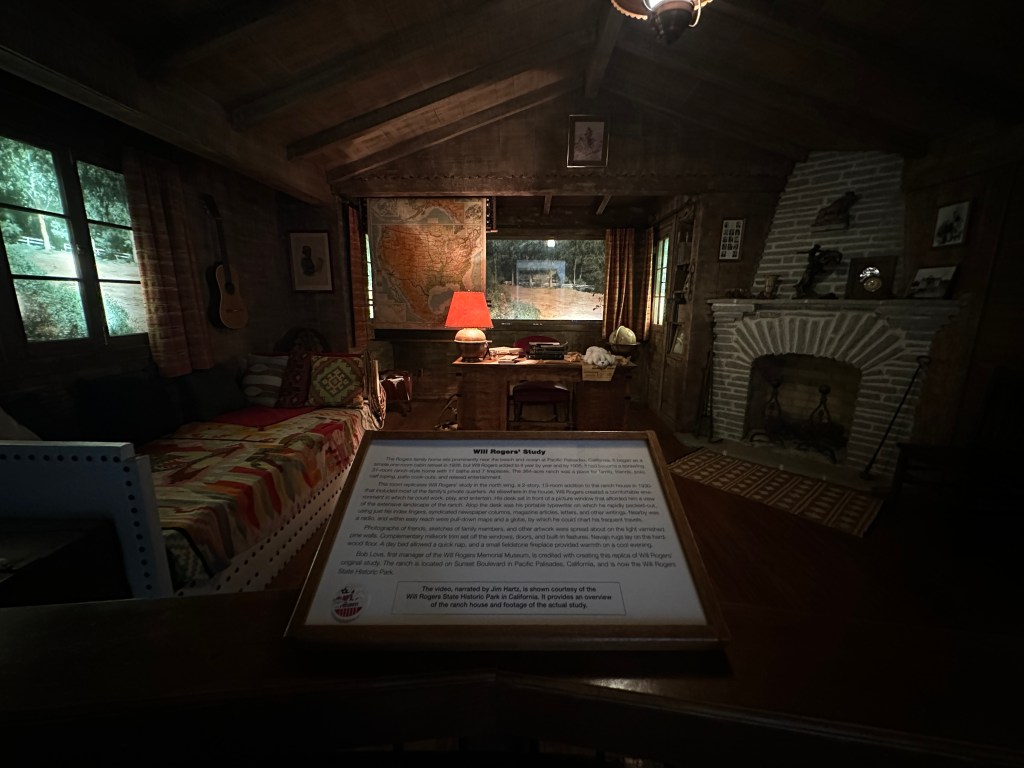 Interior view of Will Rogers' study room, featuring a rustic decor, wooden furniture, a fireplace, a desk with documents, a vintage map of the United States on the wall, and cozy seating with a quilt.
