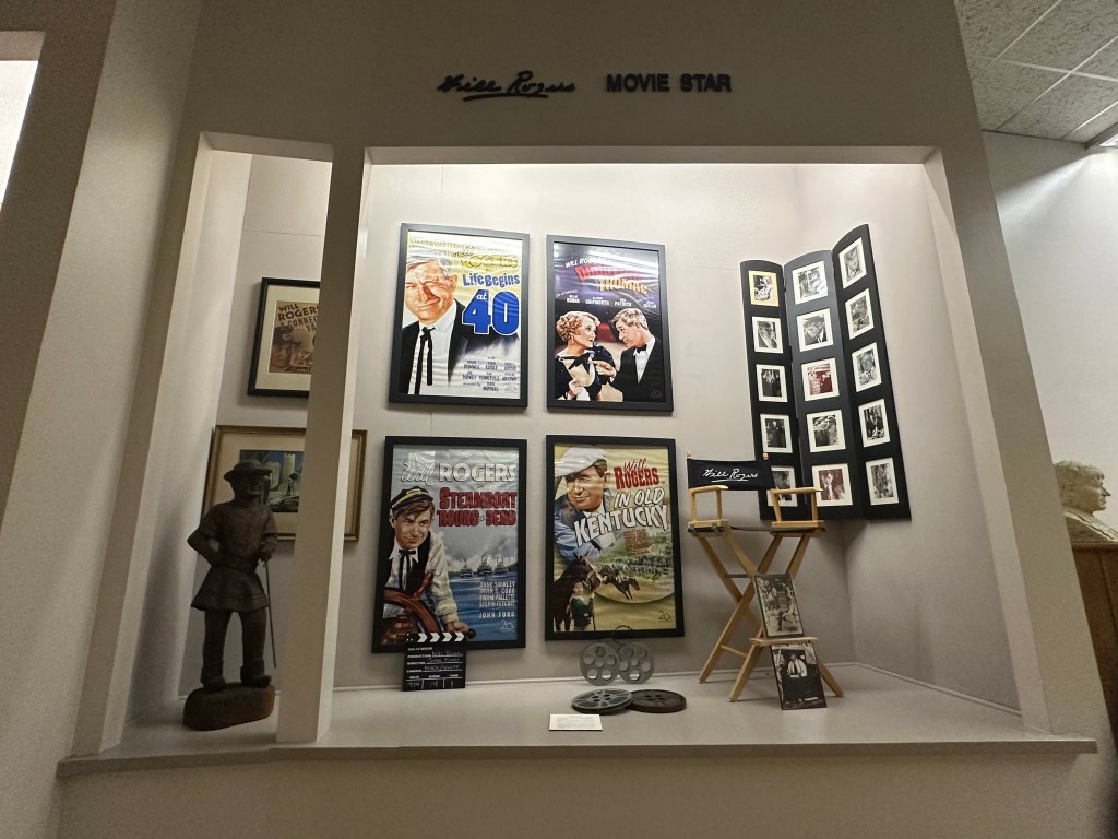 Display of movie posters and memorabilia honoring Will Rogers, including a statue and framed photographs.
