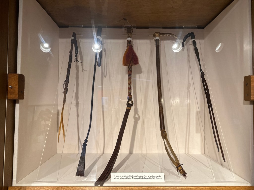A display case containing several quirts, a type of whip typically used in herding, with detailed descriptions. The quirts are from the collection of Will Rogers.