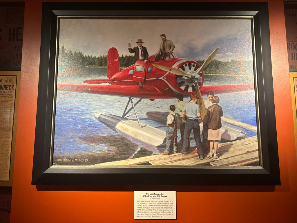 A vibrant painting titled 'The Last Farewell of Wiley Post and Will Rogers' showing a red seaplane on a lake with people gathered around, highlighting a significant moment in history.