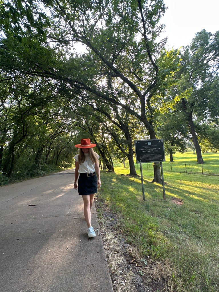 A person walking along a shaded pathway near a historic marker for Route 66 in Arcadia, Oklahoma, surrounded by lush trees.
