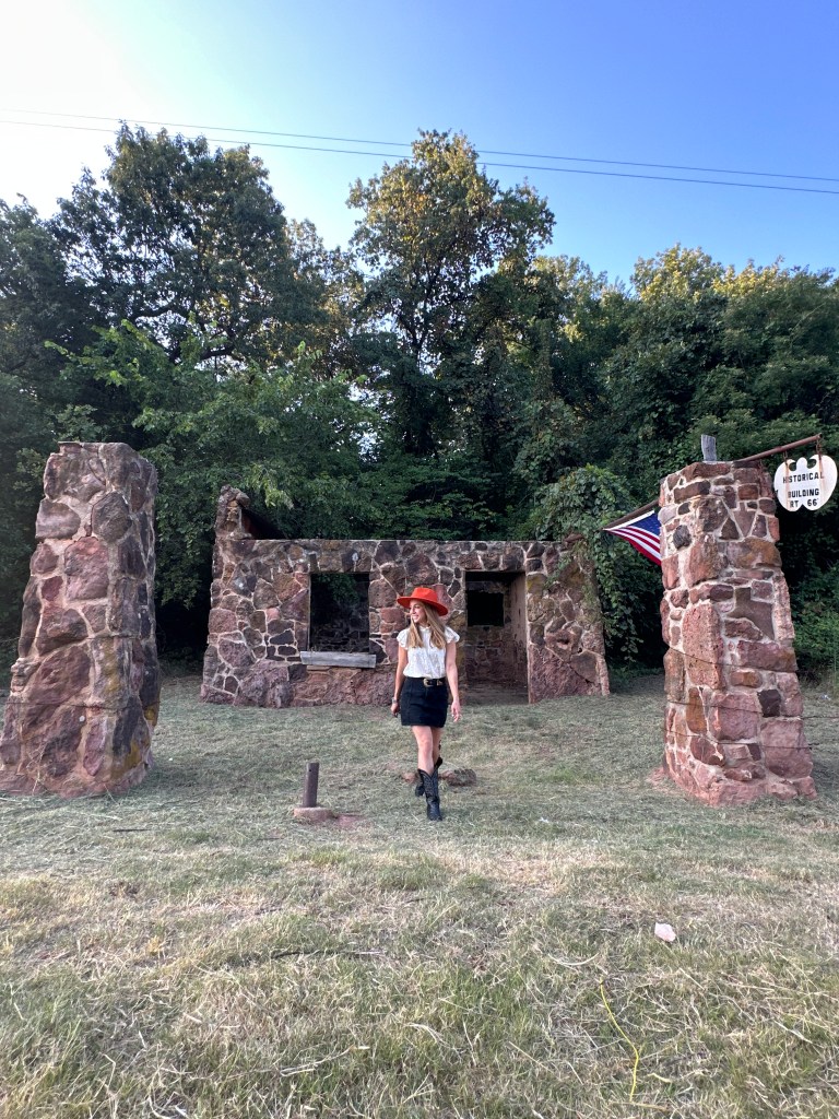 A woman in a red hat walks in front of a vintage stone gas station with two pillars and a sign indicating it is a historical building, surrounded by trees and grass.