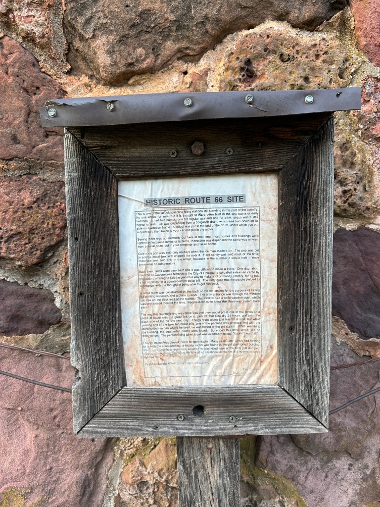 A weathered sign detailing the history of a historic gas station located along Route 66, mounted on a rustic wooden frame against a stone wall.