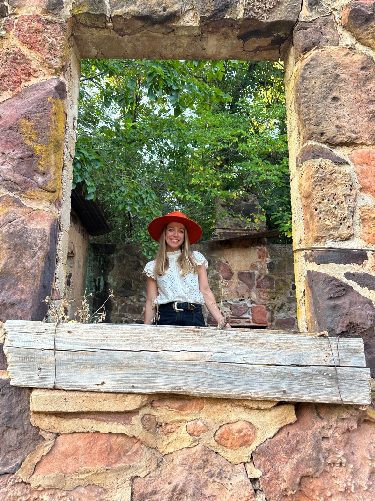 A woman standing in a window frame of a historic stone building surrounded by greenery.