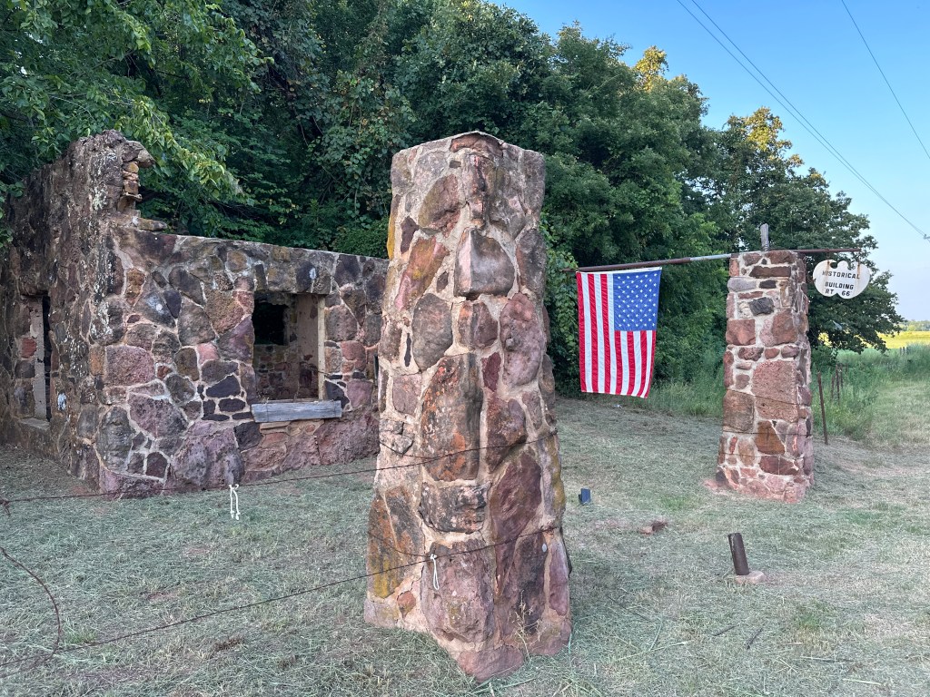 Historic stone gas station ruins along Route 66, featuring two stone pillars and an American flag.