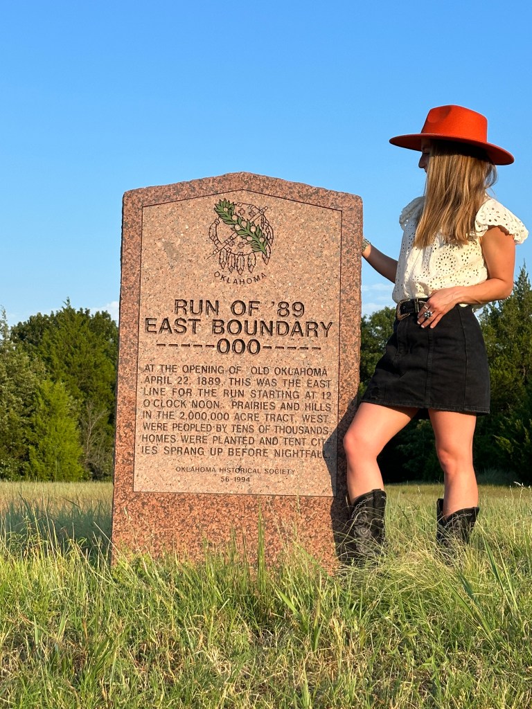A woman in a white blouse, black skirt, and red hat stands next to a historical marker titled "Run of '89 East Boundary" which commemorates the opening of Oklahoma in 1889.