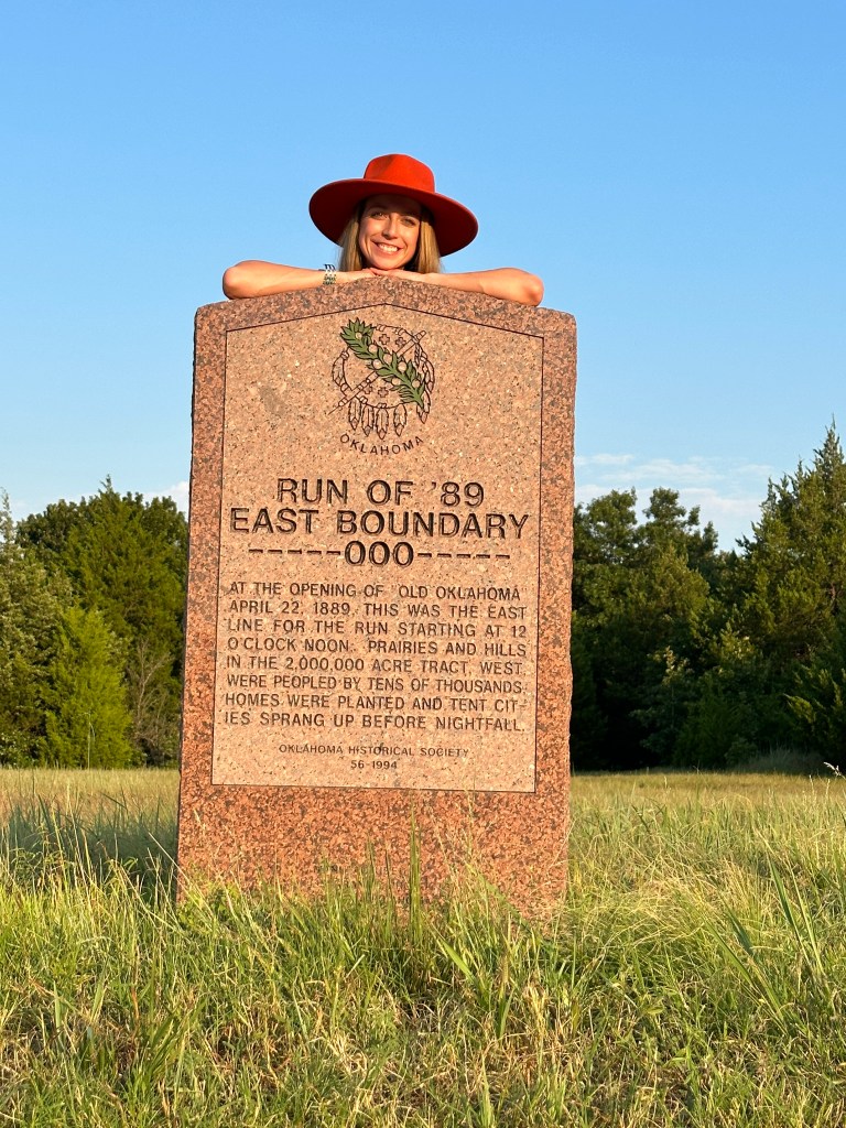 A smiling woman in a wide-brimmed orange hat is standing behind a historical marker detailing the eastern boundary line of the 1889 Land Run in Oklahoma, surrounded by greenery.