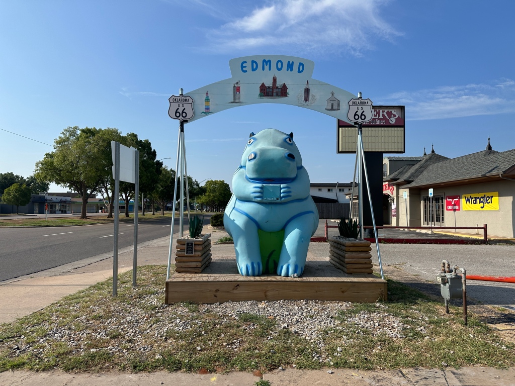 A giant blue hippo statue sits on the side of historic Route 66 in Edmond, Oklahoma, under a sign that reads 'Edmond'.