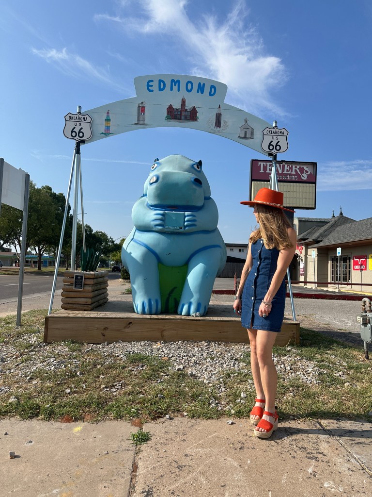 A woman stands next to a giant blue hippo statue on Route 66 in Edmond, Oklahoma. The statue is flanked by a sign that reads 'EDMOND' above it.