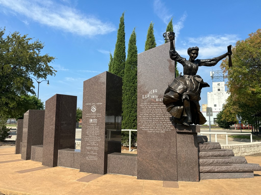 A bronze sculpture of Nannita R.H. 'Kentucky' Daisey leaping from a historical marker, surrounded by stone pillars with inscriptions about her life and achievements, set in a park in Edmond, Oklahoma.