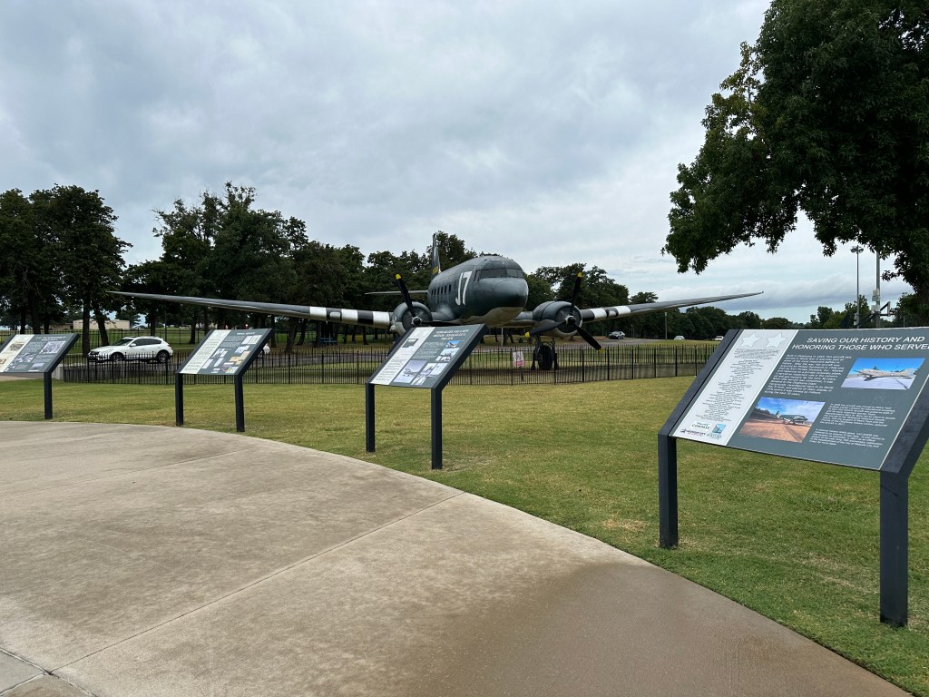 A vintage aircraft displayed outdoors, surrounded by informational signs on grassy grounds, with trees and a cloudy sky in the background.