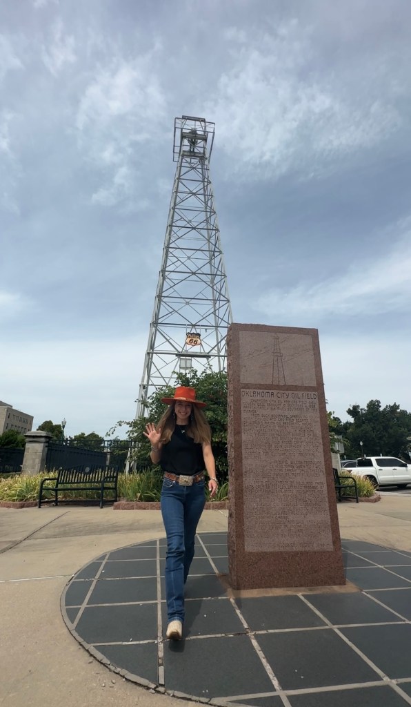 A person walking in front of a historical marker for the Oklahoma City Oil Field, with an oil rig in the background.