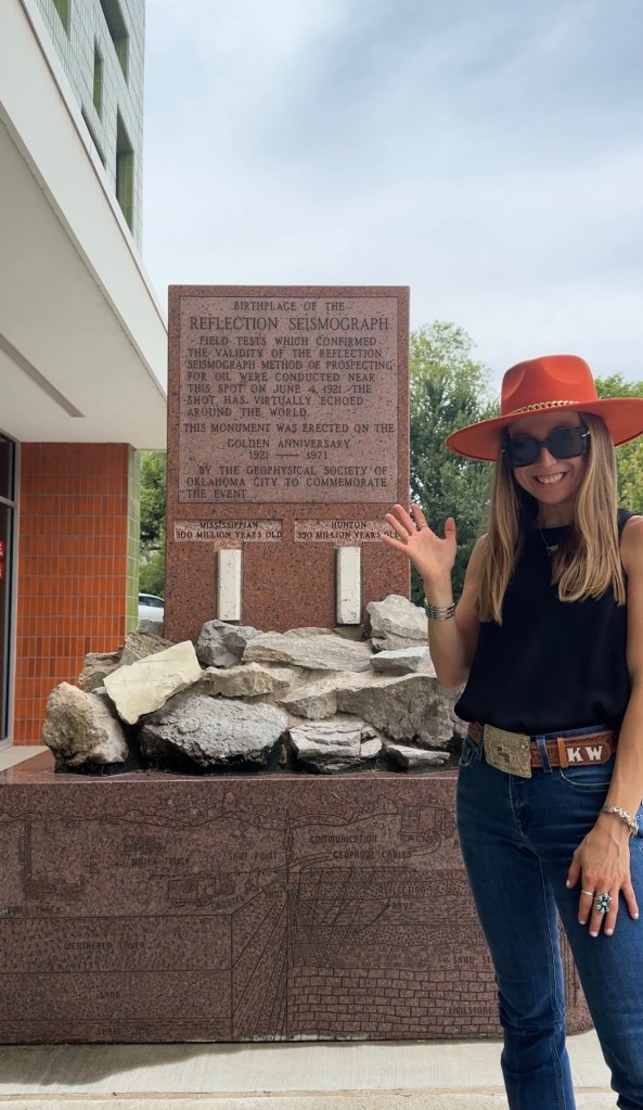 A person stands beside a historic marker at the Belle Isle Library in Oklahoma, commemorating the birthplace of the Reflection Seismograph. The marker includes text detailing the significance of the reflection seismograph method for oil prospecting, with a background of natural stones.