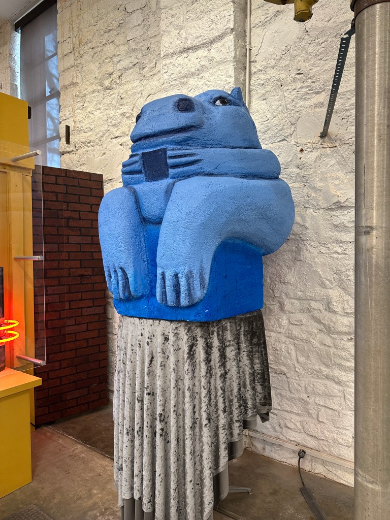 A giant blue hippo sculpture sitting with a textured gray skirt in an exhibition space, surrounded by brick and yellow display elements.