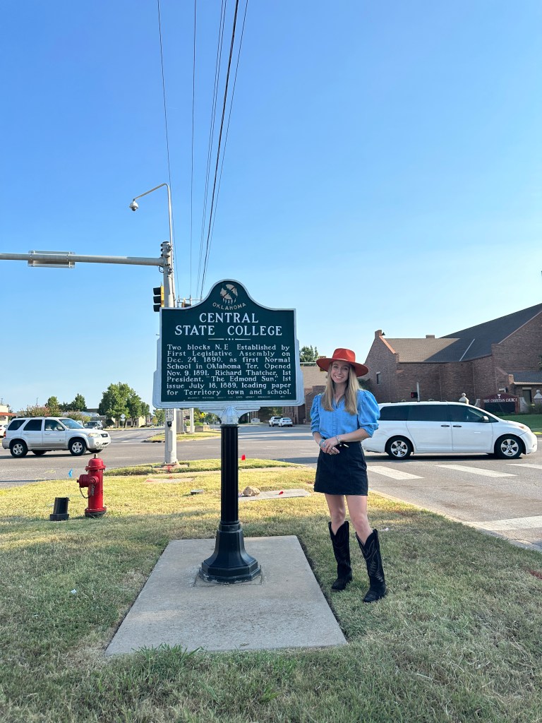 A person stands next to a historic marker for Central State College in Edmond, Oklahoma, against a clear blue sky.