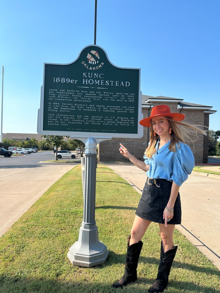A person pointing at the Kunc 1889er Homestead historic marker in Edmond, Oklahoma, wearing a blue blouse, black skirt, and cowboy boots, with a red hat.