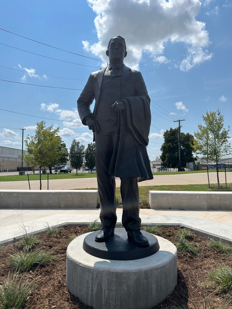 A bronze statue of a man in a suit standing confidently, holding a coat, set against a blue sky with clouds.