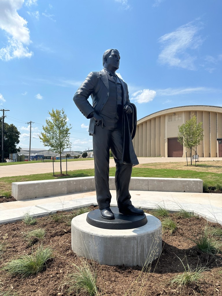 A black statue of a well-dressed man standing confidently on a circular pedestal, surrounded by small plants, with a building and blue sky in the background.