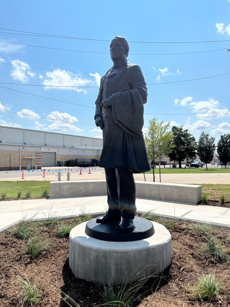 A bronze statue of a man in formal attire standing proudly on a round pedestal, with green landscaping and a clear blue sky in the background.