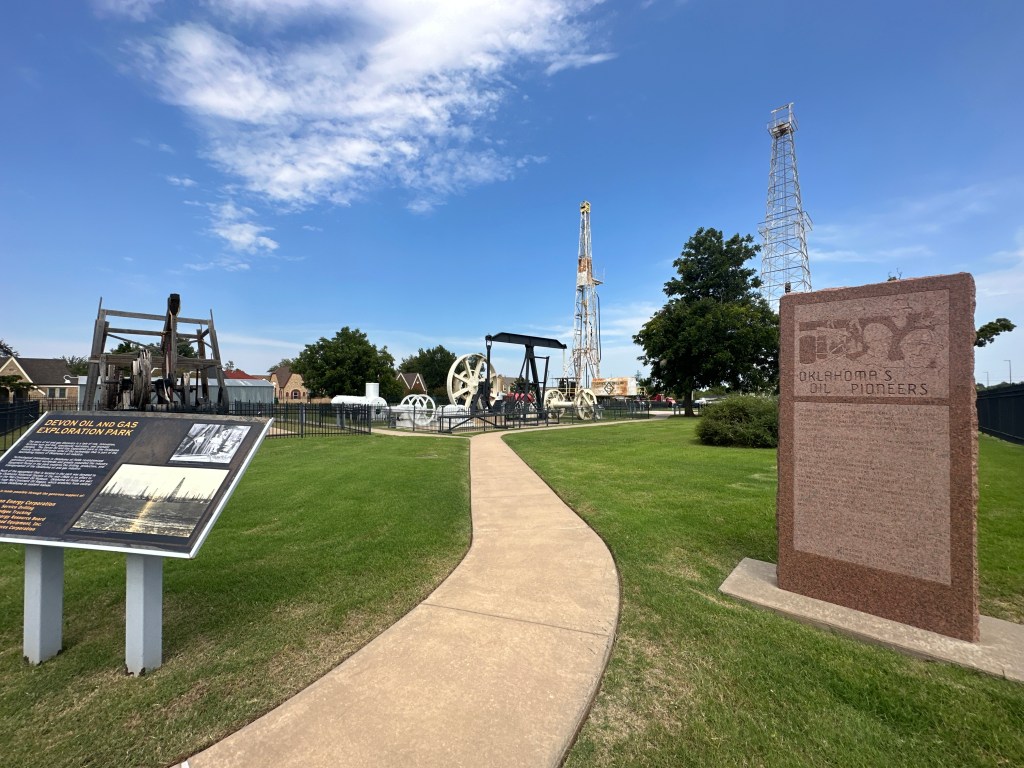 View of the Oklahoma History Center Museum's oil discovery park featuring a historic marker for Oklahoma's Oil Pioneers, with oil drilling equipment in the background and a clear blue sky.