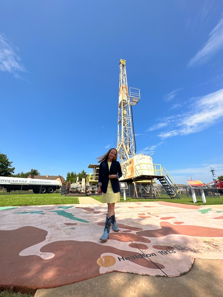 A person stands on a colorful map in front of an oil drilling rig at the Oklahoma History Center Museum, under a clear blue sky.