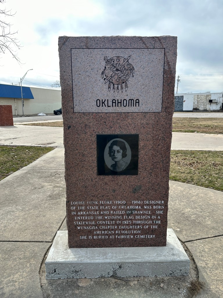 A granite monument dedicated to Louise Funk Fluke, the designer of the Oklahoma state flag, featuring her portrait and an inscription about her life and achievements.
