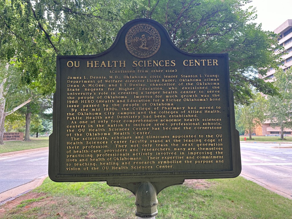 Historic marker for OU Health Sciences Center, located in a green area with trees in the background, displaying text about the university's history and mission.