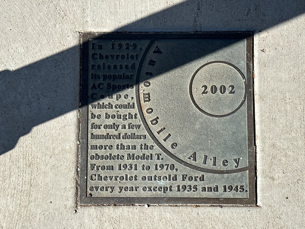 A historic marker plaque in the sidewalk, detailing the 1929 Chevrolet AC Sports Coupe and its sales history, located in Automobile Alley.