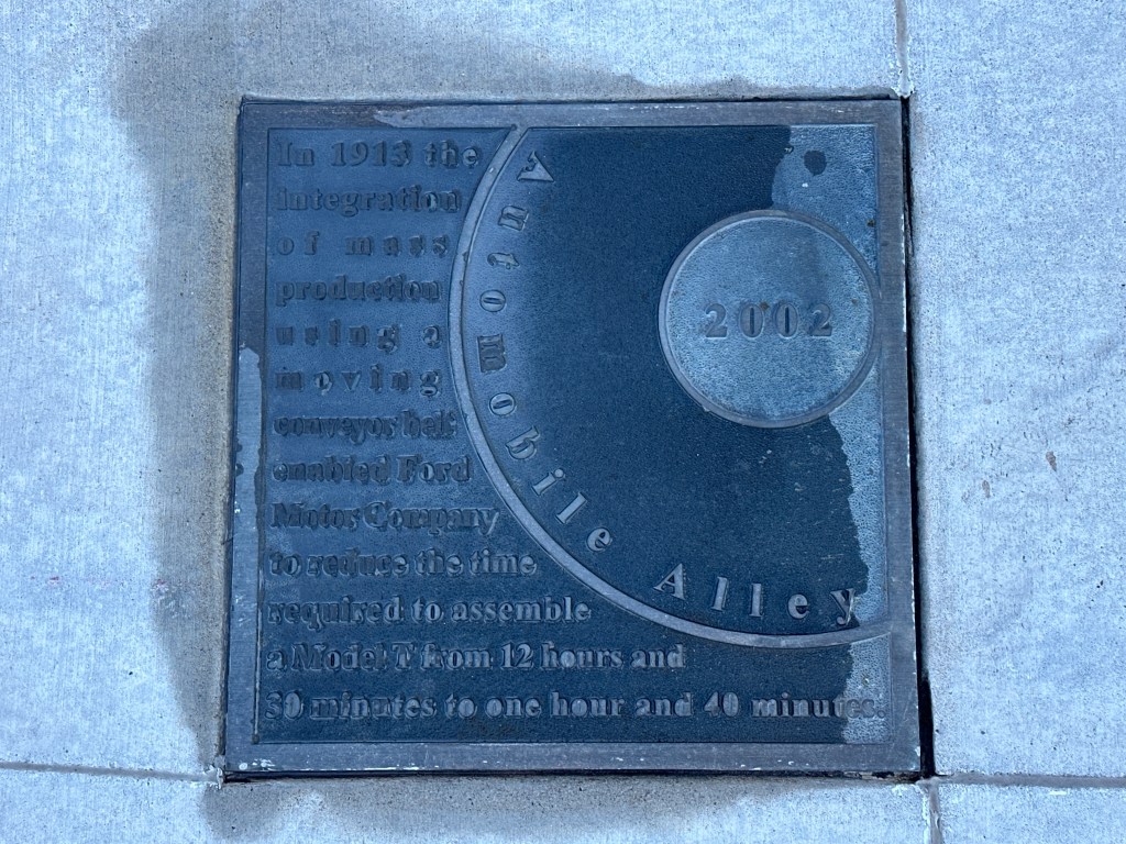 Historic marker plaque on the sidewalk in Automobile Alley, Oklahoma City, detailing the impact of a moving conveyor belt on automobile production in 1913.