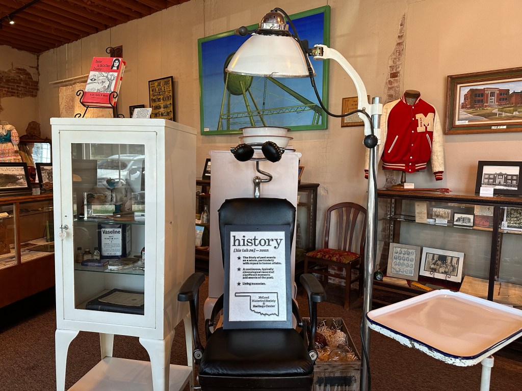 Inside the McLoud Historical Society Museum, showcasing a vintage barber chair with a sign stating 'history', along with various historical items in glass display cases and a red letterman jacket in the background.