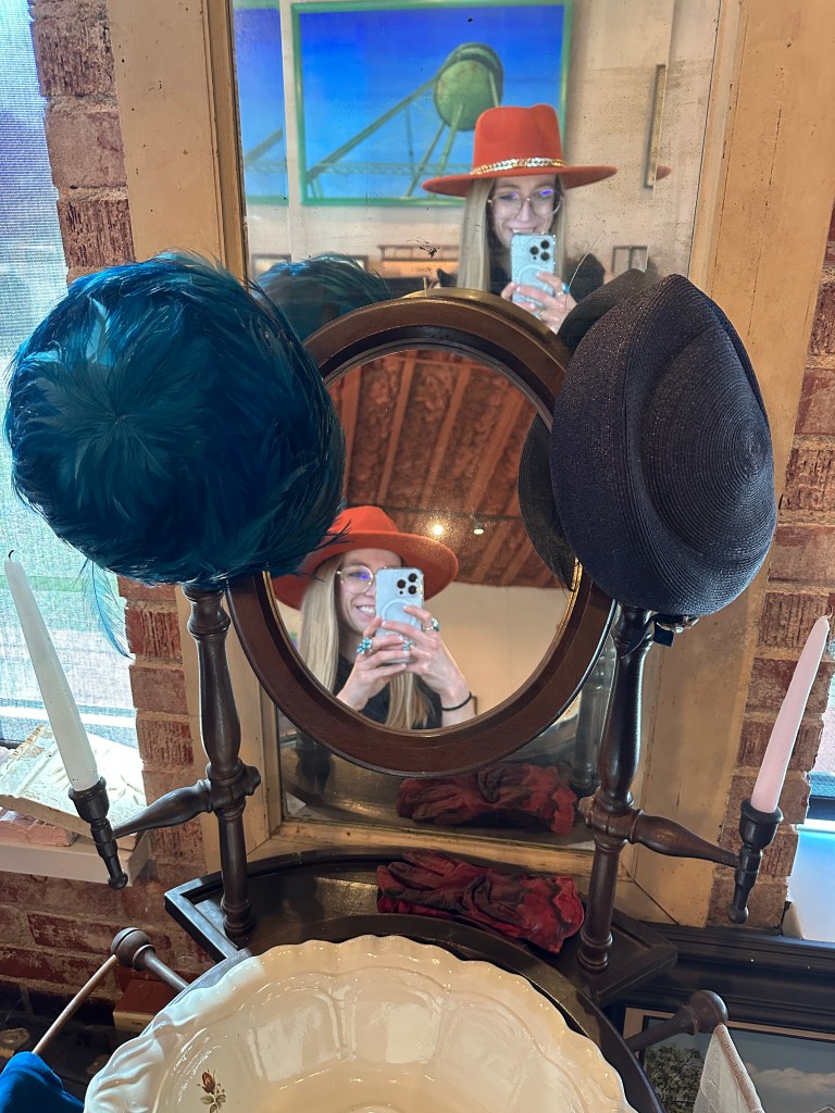 A reflection in a vintage mirror showcasing a collection of hats, including a bright red hat with a sparkly band and a blue feathered hat, with additional decor elements like candles and a bowl.